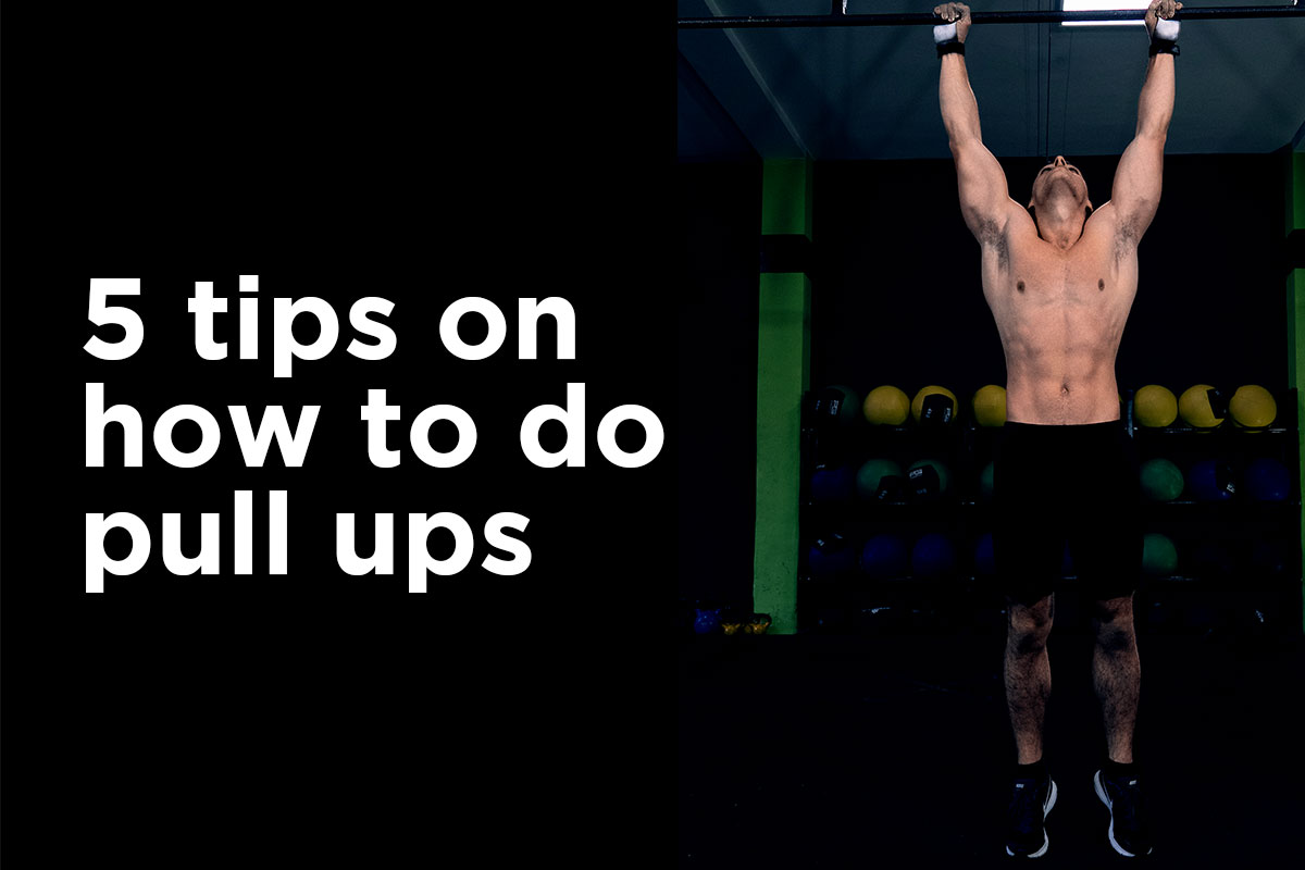 ''5 tips on how to do pull ups'' written on the left side, on a black backgorund. On a right side, a ripped man in training shorts and sneakers standing in a gym (background darkened, but gym equipment visible). He is grabbing a bar above his head, hand slightly than shoulder width apart, his gaze facing upwards towards the bar, his toes touching the floor.