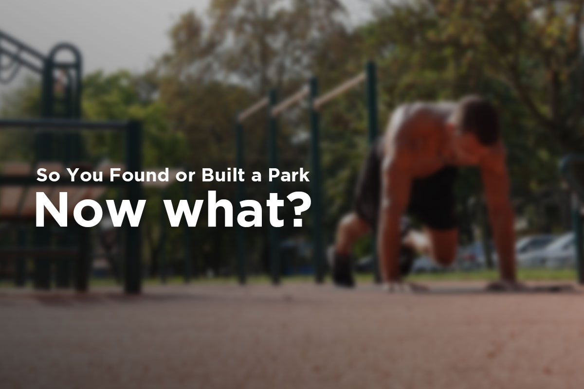 A picture of a man doing mountain climbers (push up position, the knee of the left leg is reaching inbetween arms, right leg straightened behind him) in a workout park among the trees on a sunny day. Behind him, various workout bars are visible.