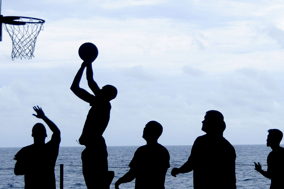 A group of men playing basketball next to the sea. Only figures are visible due to lightening.