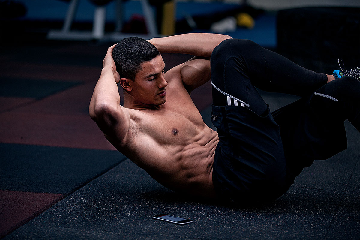 A ripped man in training shorts and snekares, lying on his back on a gym floor (gym blurred behind him), doing bicycle cruches. His  palms are behind his head. His left elbow is reaching towards his right knee, which is bent on 90-degree angle and raised from the floor 