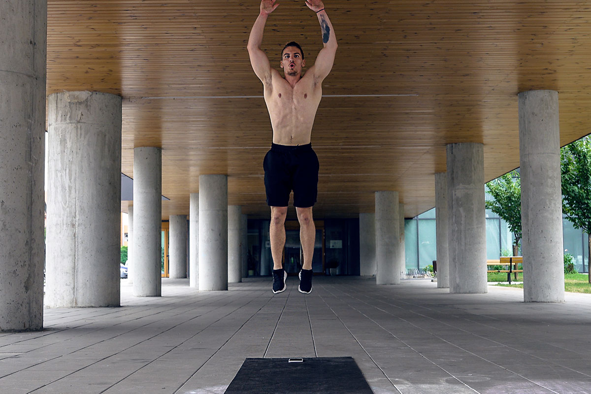 Ripped man in trainging shorts and sneakers, facing camera. He is in a yard of a big building, surrunded by a poles. The picture is taken in a mid air portion of a jump, his body fully extended, arms over head.