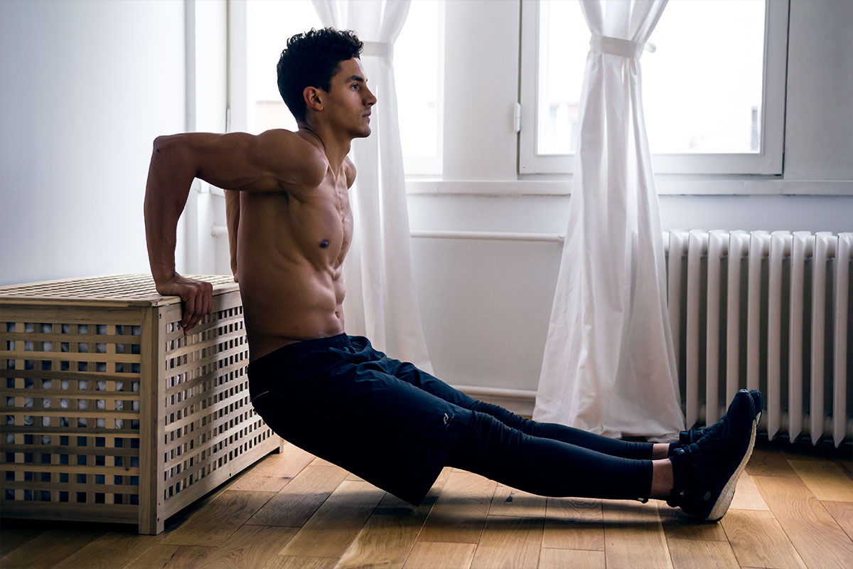 Ripped guy in a training shorts and sneakers, in a living room. He is facing backwards to a wooden bench. His palms are placed on a bench and his elbows  bent on 90 degree angle, so his upper arms are parallel to the floor. His legs are straight and in front of him, only his heels touching the floor, legs almost parallel to the floor. Exercise he is doing is named chair dips.