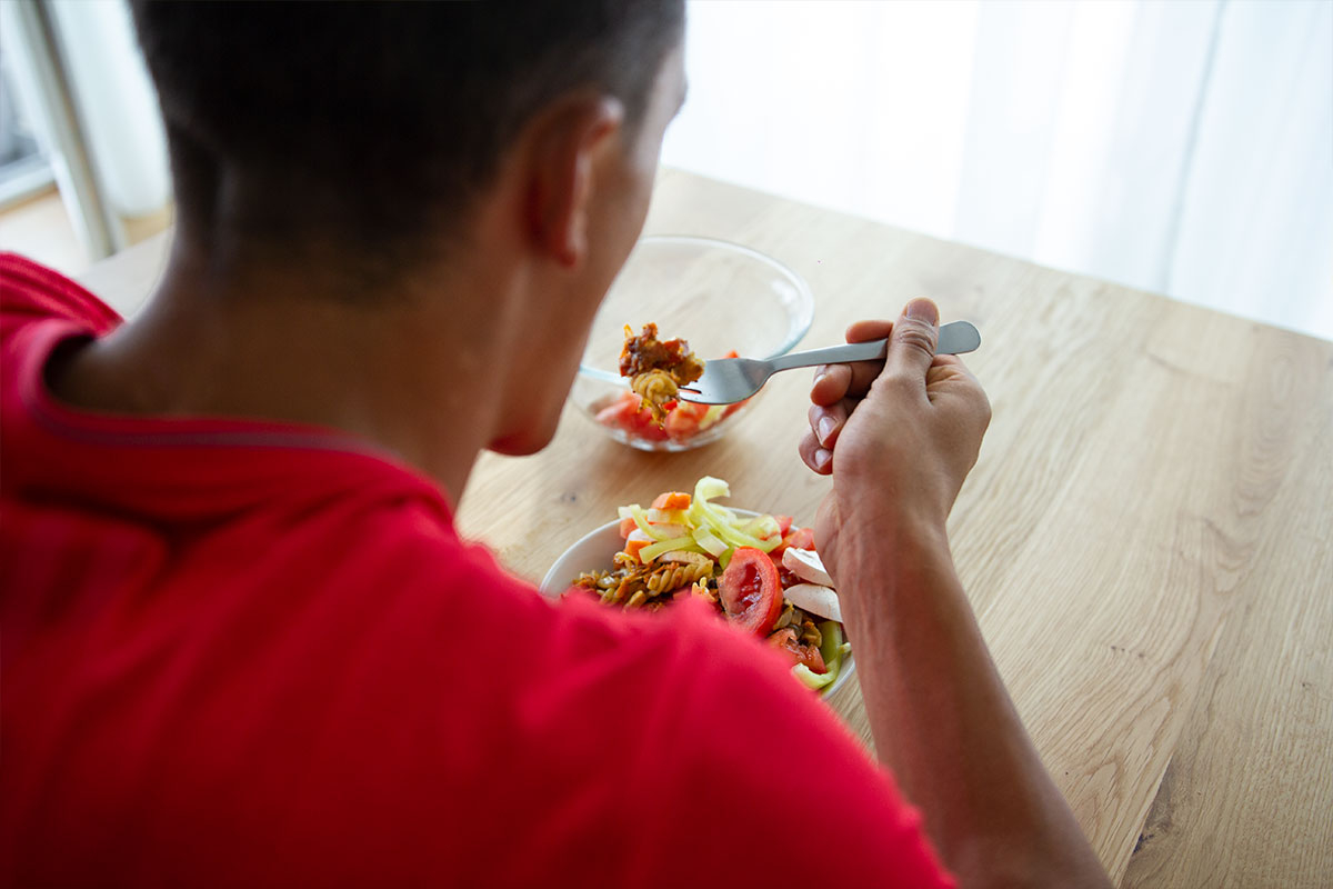 Man in red shirt, seen from behind, eating a tomato and lettuce salad.