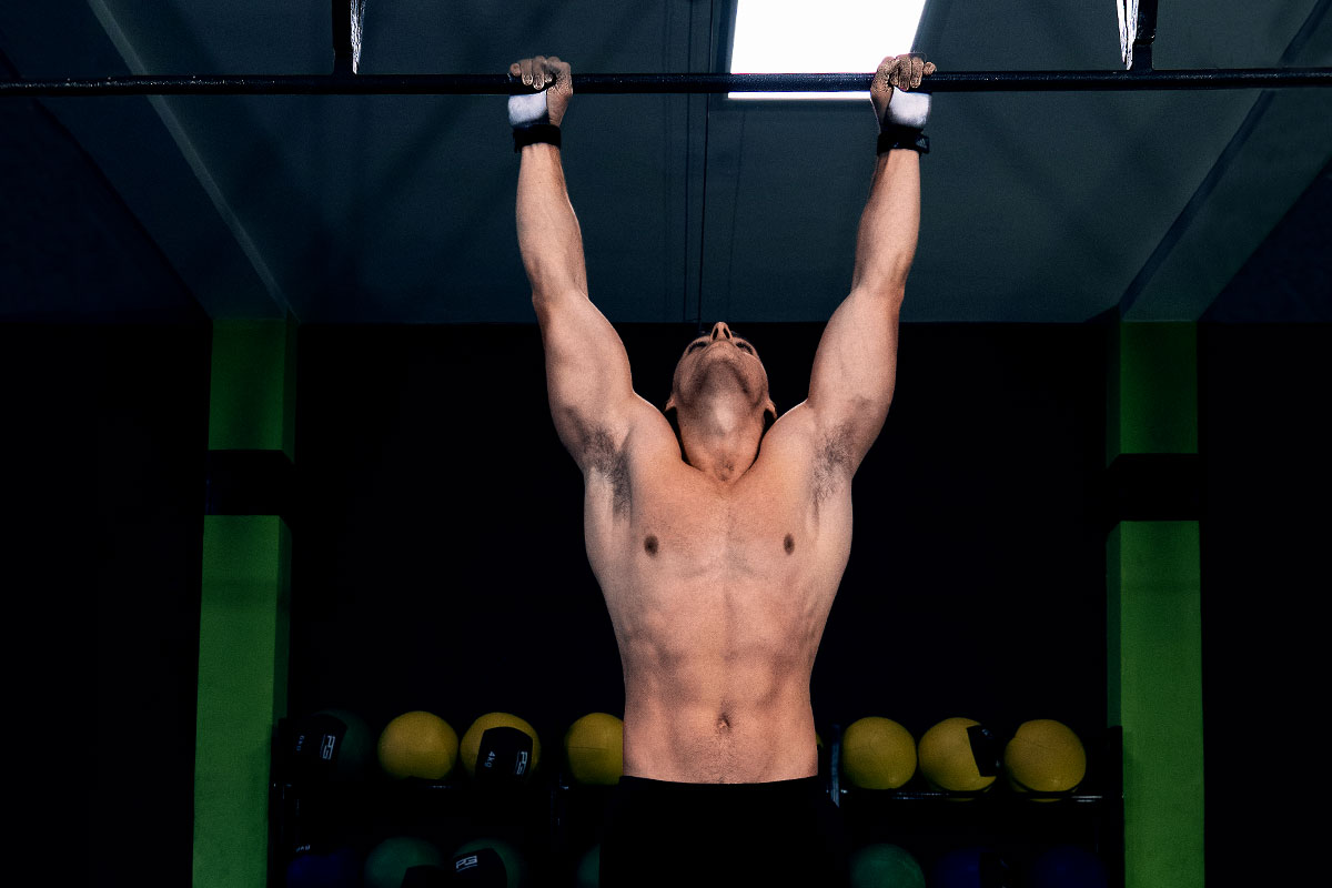 Ripped man, torzo naked, arms extender over the head, shoulder width appart, holding on to a pull-up bar, looking towards the bar.