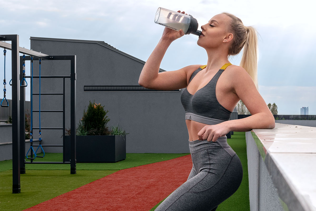 A fit blonde girl in a grey yoga pants and sports bra, standing on a rooftop with an outside gym. She is facint the camera with her left side, left elbow placed on a concrete rooftop fence, right hand holding a protein shaker filled with water, she is drinking from it. In the background, the urban scenery is visible.