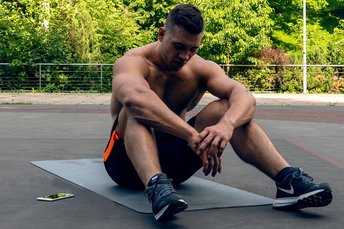 Ripped man, sitting on the mat in the playground, cellphone next to him. His elbows lying on his knees, tired from exercising.