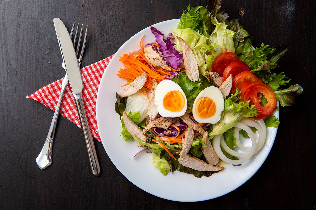 A white plate on which a salad is placed, ingredients are mixed together and one can see - lettuce, grated carrot, onions, tomatoes , and fried chicken breast pieces. On the top, two halves of hard boiled egg are placed. Next to the plate, there is a fork and a knife (on the left side). Everything is placed on a background made of dark wood.