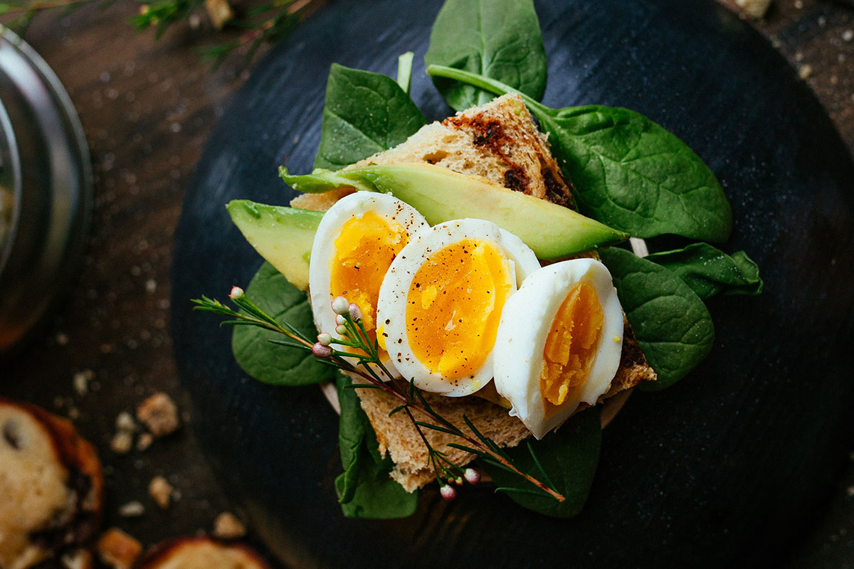 A piece of toast placed on a lamb's lettuce. On that toast, there are 3 slices of avocado, and 3 slices of hard boiled egg. The toast is placed on a black plate, on a dark wooden table.