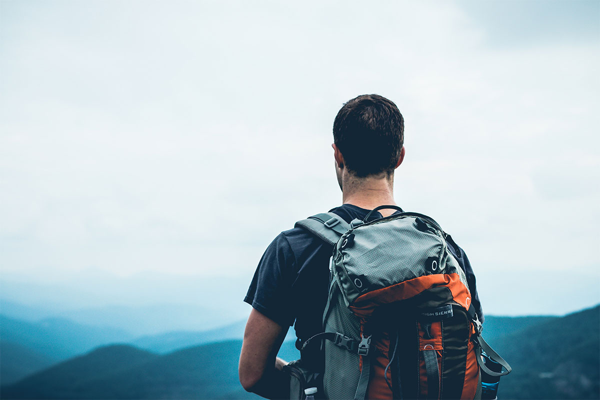 A man standing on a mountain top, facing camera backward, wearing a hiking backpack, gazing at the distance. Mountain range visible in front of him, covered in a light fog.