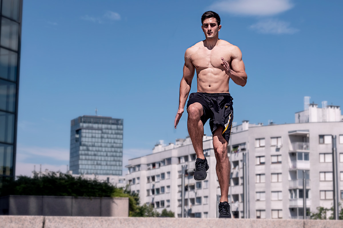 A ripped man, wearing training shorts and sneakers, torso naked, facing camera front. He is doing high knees exercise: his right knee is lifted in a 90 degrees, and his left arm bend on the elbow on 90 degrees. Other arm ang leg are straight, left foot on the floor. Behind him, urban landscape is visible.