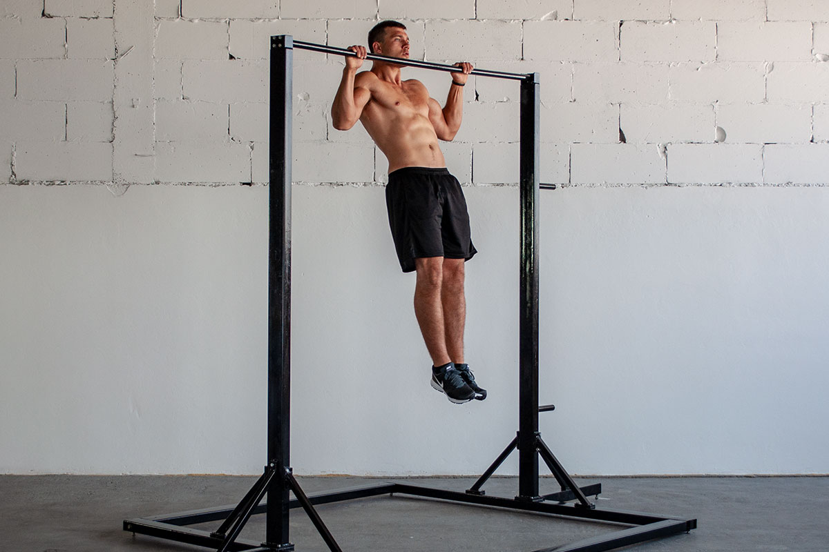 A ripped man in training shorts and sneakers, standing in a vast room, white wall visible behing him. There is a free standing pull up bar in the room, and he is holding a pull up hold position on that bar (arms holding for a bar, slighltly more than shoulder width apart, elbows bent, he is pulling his chin over the bar). He is facing the camera.