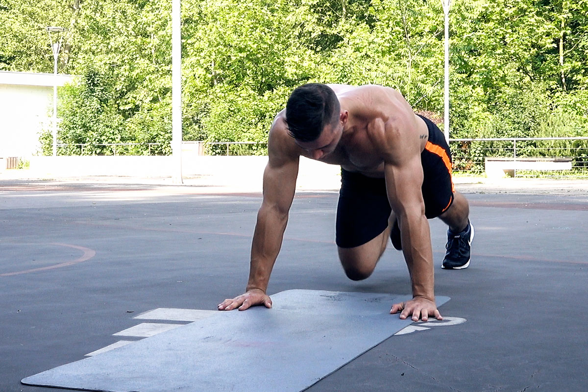 Ripped man in a training shorts and sneakers on a playgroung, sorrounded by a trees. He is facing the floor, standing on his palms, hands fully extended. His left leg is extended behind him and his toes touching the floor. His righ leg is bent in the knee and his knee is reaching for the inside of his left elbow. The exercise he is doing is called the mountain climbers.