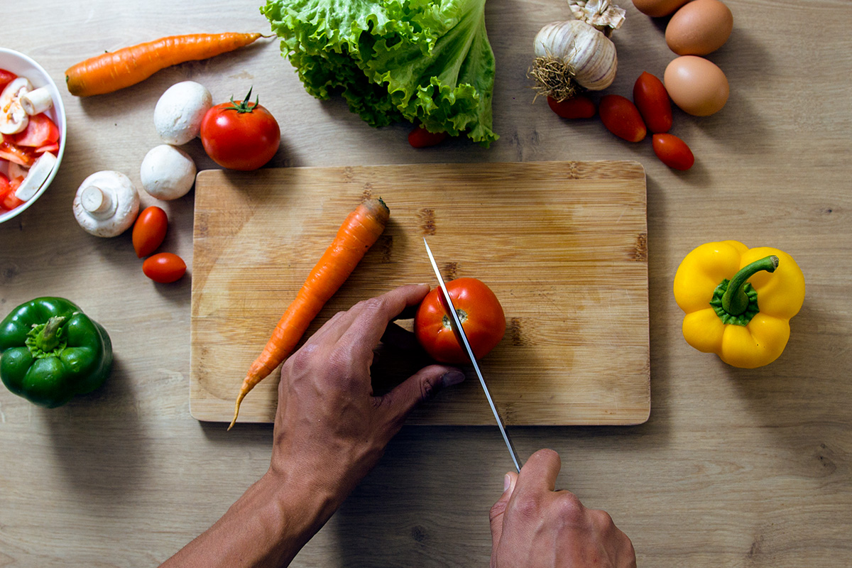 Wooden table with wooden chopping board, vegetables and eggs lying aroun it, two male hands seen holding the knife and cutting tomatoe.