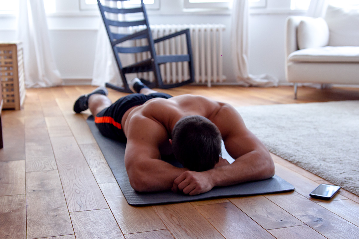 Fit man in sports equipment lying on a yoga mat on his belly, head resting on the hands, facing the mat, phone by his side