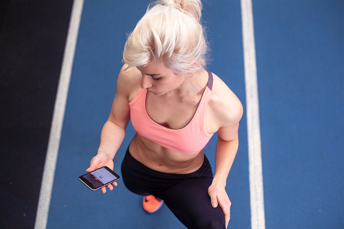 A blond fit girl, camera is looking at her from above, she is wearing pink sports bra and sneakers, as well as black joga pants. In her right hand, she is holding a cellphone. She is standing on a blue running track.