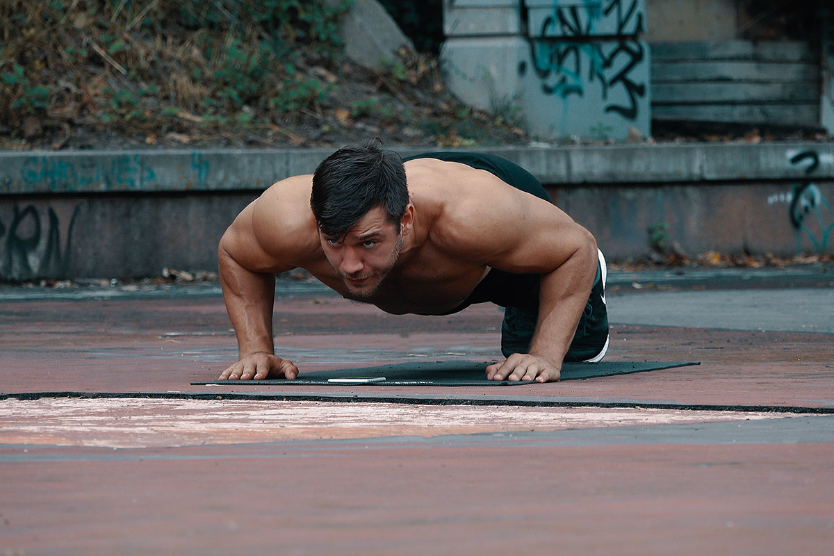 ripped man in training shorts and sneakers, facing the camera, in a push up hold position. Palms and toes toucing the floor, back straight, elbows bent in 90 degree angle and slightly away from torso.