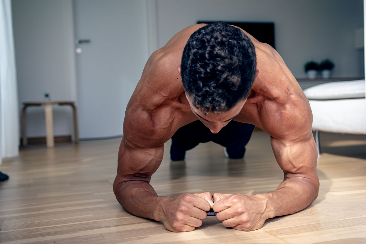 Ripped man i a training shorts in a livingroom of the apartment decorated in white and grey. He is facing camera in a plank position - hiy body straight and parallel to the flor, his toes and forearms are touching the floor. Shoulder and arm muscles flexed. 