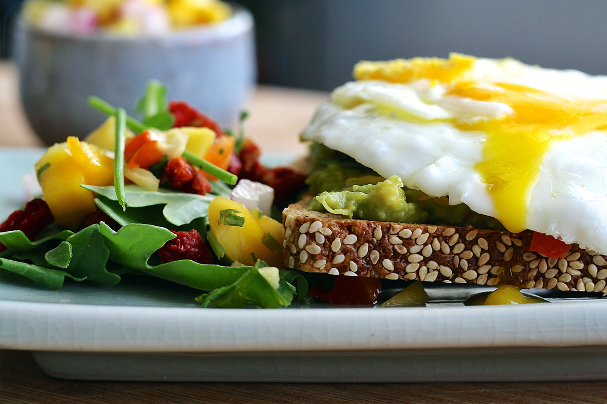 A plate with food on it. On the left, there is a mango and lettuce salat, on the right a slice of brown bread  with mashed avocado and fried egg on the top. 