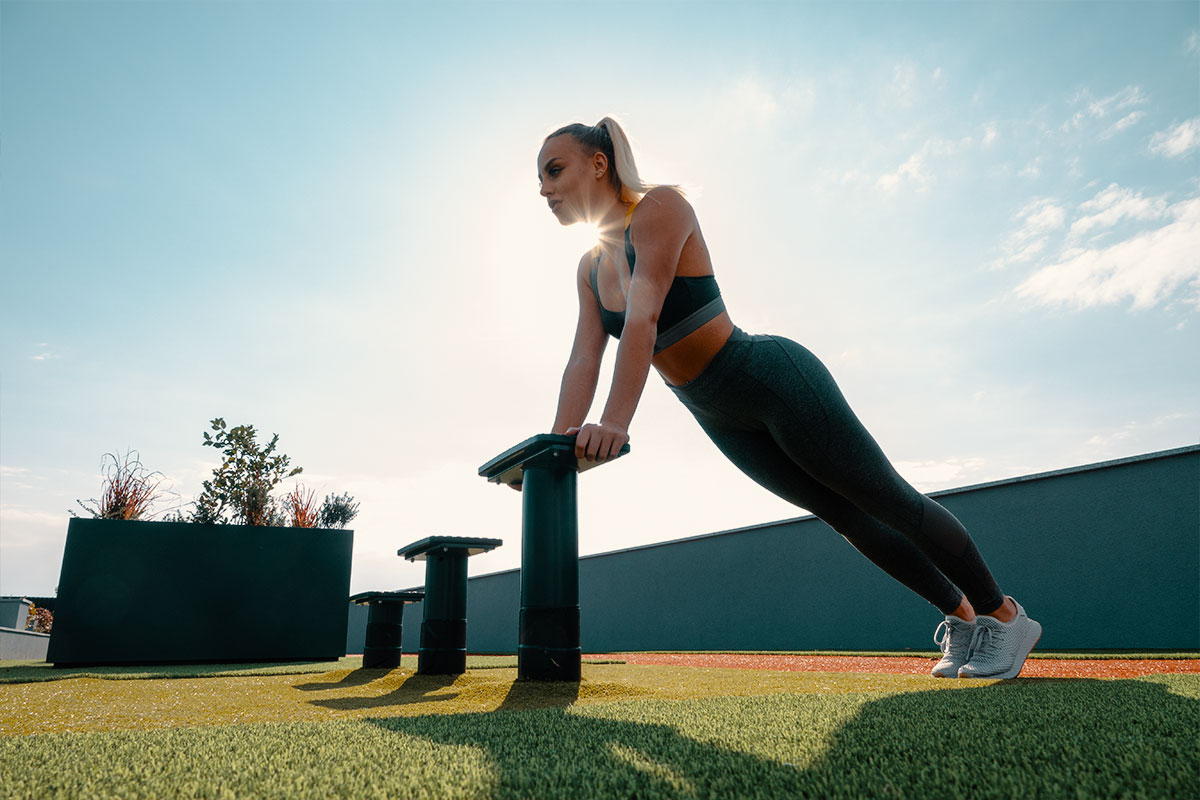 A blong girl, wearing grey yoga pants, sports bra and snekares, She is in an outdoor space, what seems to be like a rooftop with a grass surface and atheltic trail on the edge, near the fence. There are three small benches, and she is leaning with her hands on one of those benches, standing in an incline push up position (hand straight and inclined on the bench, body staight).