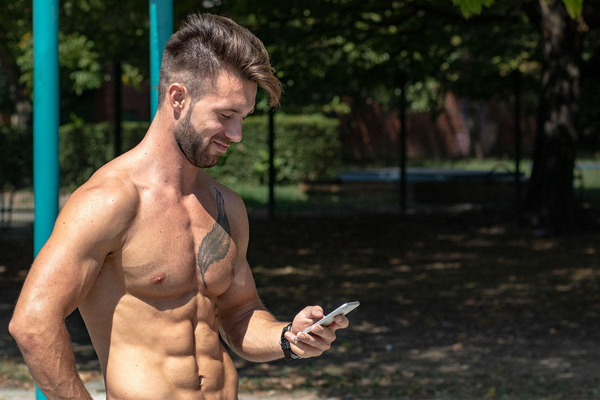 A ripped man, only upper body shown on the picture, standin in a workout park in the nature, holding and looking at the mobile phone in his right hand.