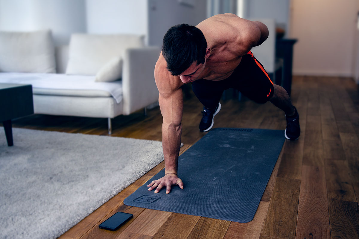 A ripped man in training shorts and sneakers, faing the camera forwards, standing in a one-arm-push-up position. Left arm bent behind his back, palm of the straightened right arm and toes touching the floor, back straight. 