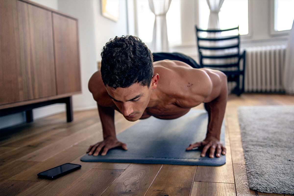 Ripped man in an apartment livingroom, wearing trainging shorts. He is facing the floor, his palms and toes are on the floor, his body straight and parallel to the floor, his elbows bent. The exercise he is doing is called push up hold.