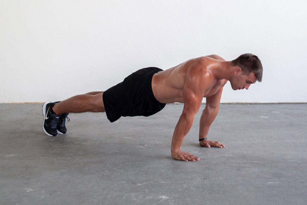 Ripped man in trainging shorts and sneakers, standing in a plank position (toes and palms touching the floor, body and arms straight, face facing the floor. He is facing the camera with his right side.