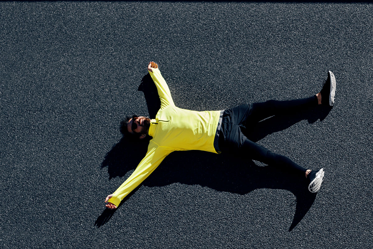 A man in a yellow long-sleeved sports shirt, black long sports tights, and black sneakers, lying on a playground floor, arms and legs extended in a starfish position.