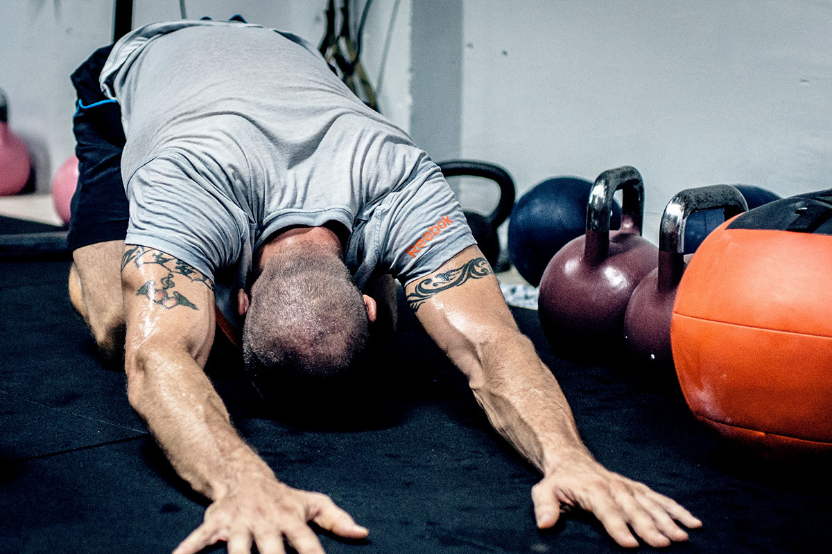 A muscular man, wearing black shorts and a grey t-shirt, on his knees, sitting on his heels, forehead touching the floor, arms extended in front of his body (yoga - child pose). Various kinds of weights can be seen around him.