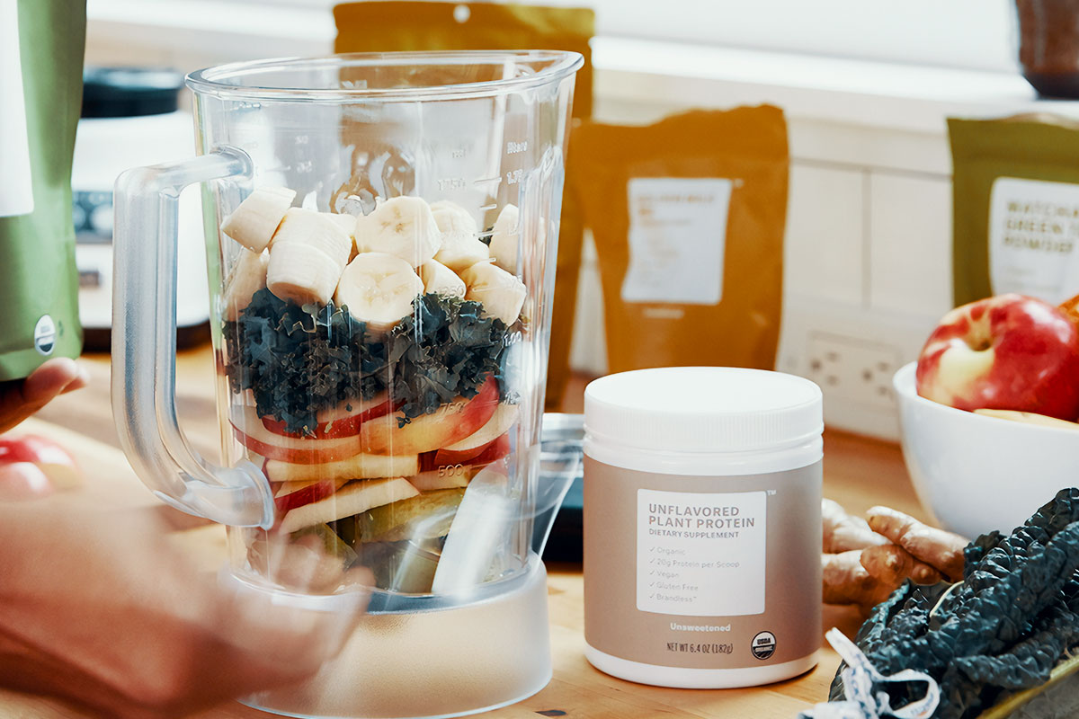 A blender bowl with sliced fruit lined up in it. Apples, pears, kale, and banana are visible in a bowl. Next to the bowl, a white containter with golden wrap on which is written ''Unflavored plan protein'' is placed. Around the bowol, various foods are visible, such as kale, apples in a bowl, ginger, and bags with various spices.