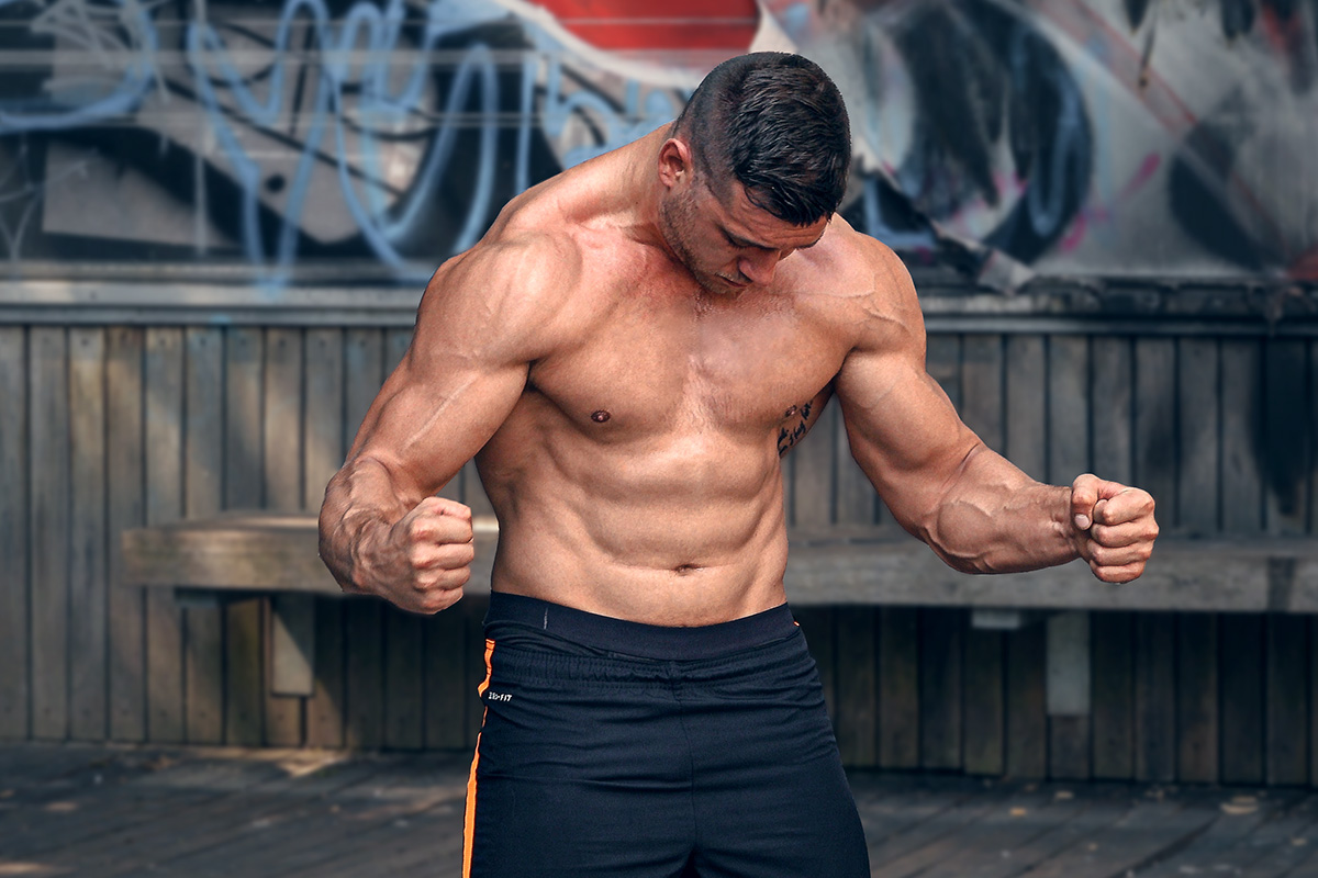 Ripped man, graffiti wall in the backgroud, torso naked, flexing his arms and abs, looking at his abs.