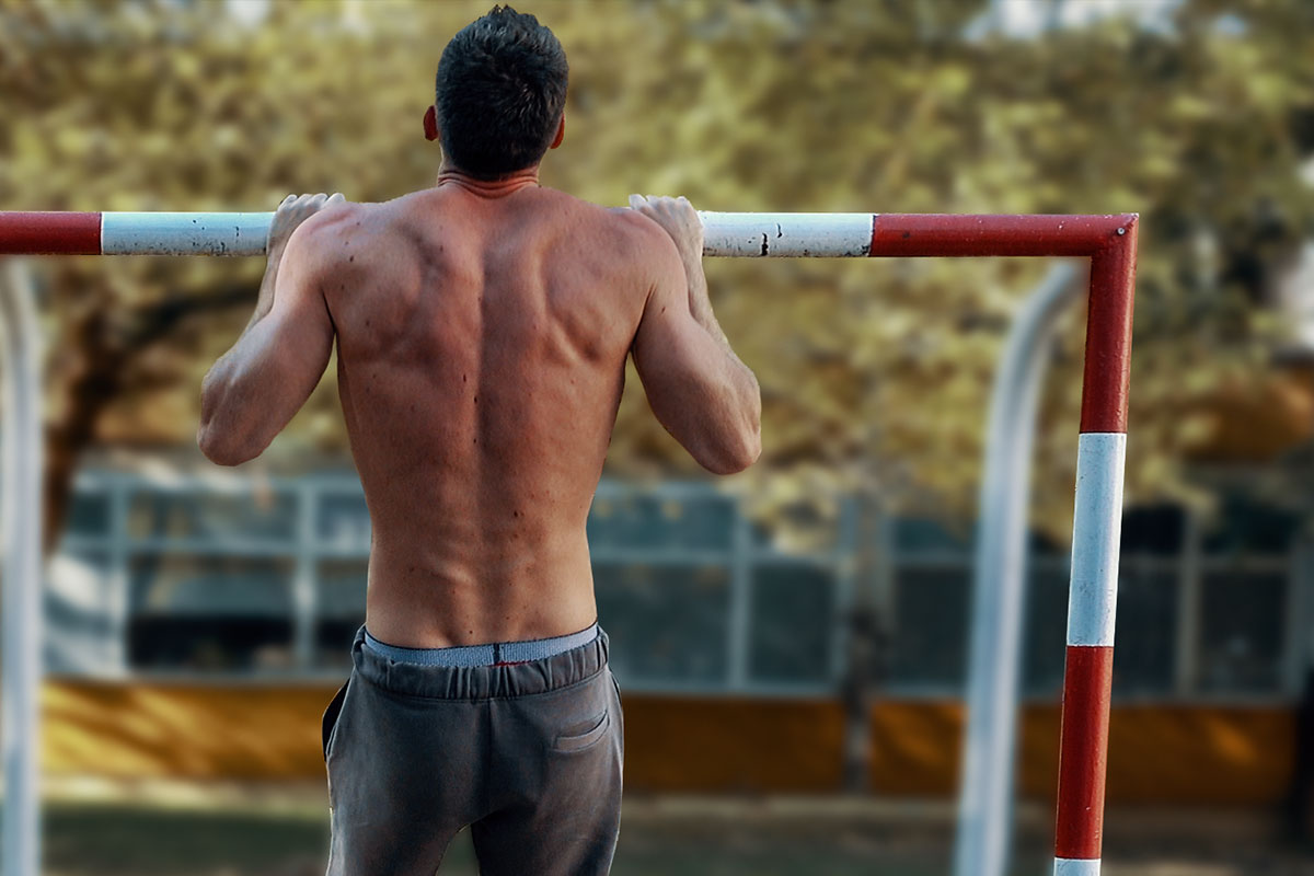 a ripped man in training shorts, ouside in a park. He is stading in a pull up hold position using an empy frame of soccer goal as a pull up bar. Pull up hold position: (arms holding for a bar, slighltly more than shoulder width apart, elbows bent, he is pulling his chin over the bar).