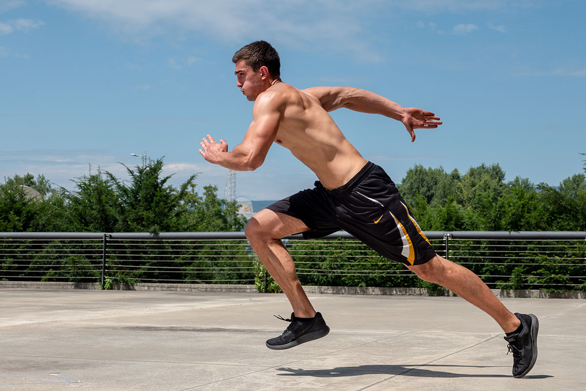 A ripped man, wearing training shorts and sneakers, torsko naked, sprinting on the concrete floor. Behind him, ther is a  fence visible, as if he is on an elevated surface. Behind the fence, one can see tops of the trees and some parts of urban landsacape visible throught the trees.